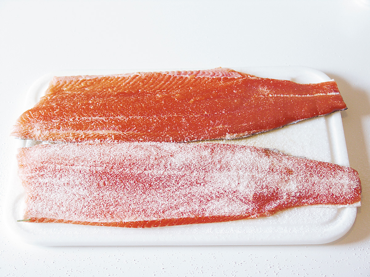 Lachs beizen: Die beiden Filets werden, Rücken an Rücken, mit der Hautseite nach unten auf ein Küchenbrett gelegt und gleichmäßig dünn mit Meersalz bestrichen. Dann kommen die Filets für ca. eine Stunde in den Kühlschrank. Nach der kleinen Pause wird eine der beiden Filethälften noch einmal kräftiger mit Meersalz bestreut. Richtwert: Streuen Sie einfach so viel Salz drauf, dass das Weiß des Salzes die rote Farbe des Fleisches fast vollständig überdeckt. Foto: BLINKER