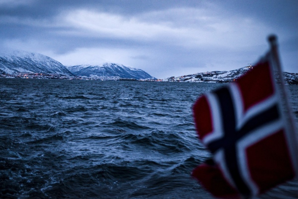 Im Vordergrund eine unscharfe norwegische Flagge aus dem Meer. Im Hintergrund Meer und am Horizont die verschneiten Berge und Lichter einer Stadt.