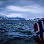 Im Vordergrund eine unscharfe norwegische Flagge aus dem Meer. Im Hintergrund Meer und am Horizont die verschneiten Berge und Lichter einer Stadt.
