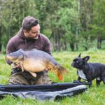 Angler hebt Karpfen aus Tragetasche und schwarzer Hund schnuppert.