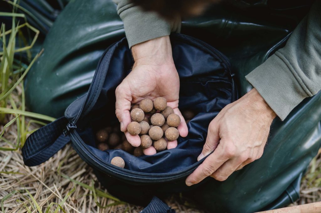 Eine Handvoll brauner Boilies über einer Art Falteimer