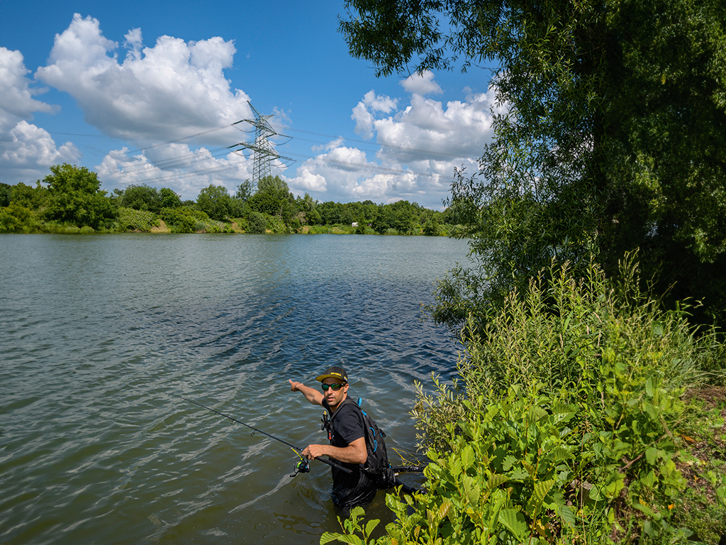 Angler am See, er zeigt auf den Schatten, den ein Baum aufs Wasser wirft